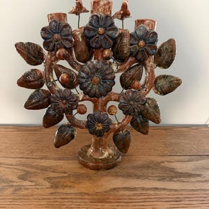 May include: A brown glazed ceramic candelabra featuring a tree-like structure adorned with dark brown sculpted flowers and leaves. The piece has three candle holders at the top and small bird-shaped figures perched on the outer branches.