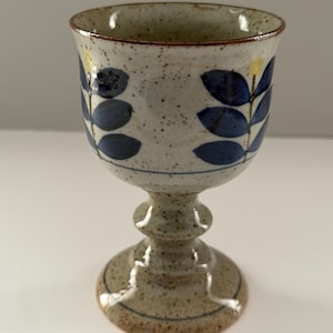 Pode incluir: Um cálice de cerâmica com acabamento bege salpicado. O cálice apresenta um padrão de folhas azul escuro e uma borda marrom. A haste tem um design decorativo em camadas. Um detalhe amarelo é visível perto da borda.