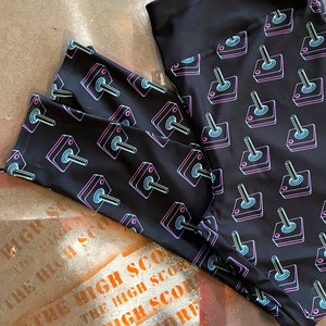 May include: Black leggings featuring a repeating pattern of neon pink and blue arcade game joysticks. The leggings have a wide, solid black waistband. The material is stretchy. The background is a distressed surface.