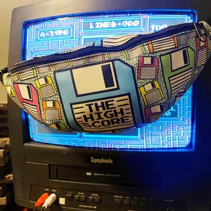 May include: A colourful fanny pack with a repeating floppy disk design in pink, yellow, blue, and green. The pack displays the text "THE HIGH SCORE" on a large blue floppy disk graphic. The pack is set against a vintage television screen.
