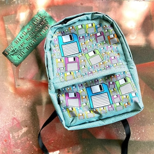 Puede incluir: Una mochila azul turquesa con un patrón de disquetes coloridos. La mochila tiene un bolsillo con cremallera en la parte delantera y correas ajustables.