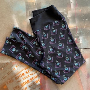 May include: Black leggings with a repeating pattern of neon pink and blue retro arcade game joysticks. The leggings have a wide, solid black waistband. The background is a distressed, multi-coloured surface.