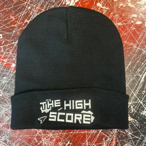Może przedstawiać: Czarna wełniana czapka z haftowanym białym logo z napisem "THE HIGH SCORE" z pikselowaną strzałką wskazującą w lewo.