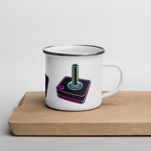 May include: White enamel mug with a retro-style joystick design in neon blue and pink. The joystick is on a black base with a white button.