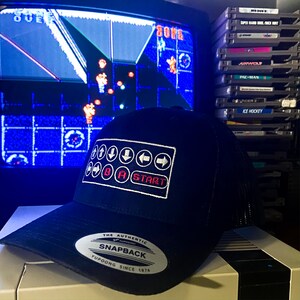Könnte beinhalten: Schwarze Snapback-Kappe mit weißem und rotem Controller-Design. Die Kappe zeigt den Text "START" und Richtungstasten. Die Kappe liegt auf einer weißen Konsole, im Hintergrund läuft ein Retro-Videospiel.