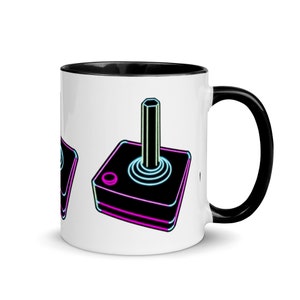 May include: White ceramic mug with a black rim and handle. The mug features a neon pink and blue joystick graphic.
