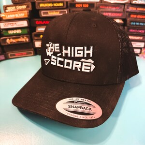 May include: Black trucker hat with the text "THE HIGH SCORE" in white. The hat has a curved brim and a mesh back. The hat also has a "SNAPBACK" tag.