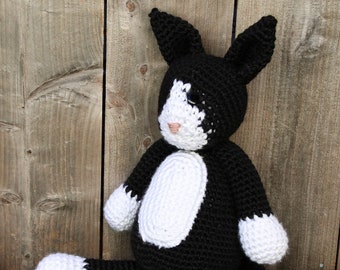 Puede incluir: Un juguete de gato de crochet negro y blanco con patas blancas y una mancha blanca en la cara. El gato está sentado en una superficie de madera.