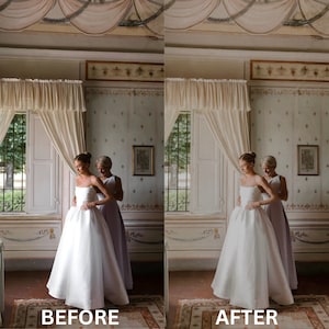 May include: A bride in a white wedding dress is being helped into the dress by a woman in a lavender dress. The bride is standing in a room with white curtains and a patterned wallpaper.