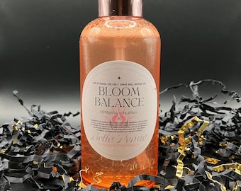May include: A clear bottle of Bloom Balance refreshing yoni spray with a rose gold cap and label. The label reads "Bloom Balance Refreshing Yoni Spray" and "Bella Penda". The bottle is surrounded by black and gold confetti.