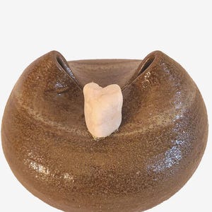May include: A brown ceramic bowl with a textured surface and two raised sides. A light pink, heart-shaped object rests in the center of the bowl. The bowl has a wide, shallow design.