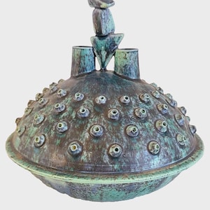 May include: A dome-shaped ceramic vessel with a mottled teal and brown glaze. The surface features a grid pattern and numerous small, raised circular protrusions. Two cylindrical openings sit at the top, topped by a stacked abstract sculpture.