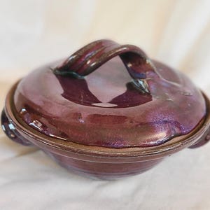 May include: A handmade, ceramic casserole dish with a lid. The dish is a deep purple color with a glossy finish and a looped handle. The rim and handle have a darker, almost black, accent. The dish has small handles on either side.