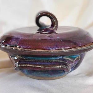 May include: A handmade ceramic bowl with a lid. The bowl is glazed in shades of purple, blue, and brown. The lid has a loop handle. The bowl's exterior features a textured design. The piece is likely used for serving or storage.