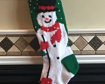 May include: A green and white knitted Christmas stocking with a snowman design. The snowman is wearing a black top hat, a red scarf, and holding a stick. The stocking has a white cuff and is decorated with white snowflakes.