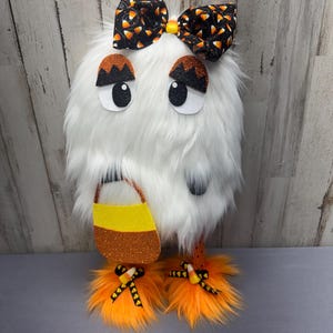 May include: A whimsical Halloween decoration featuring a white, fluffy creature with large eyes and a candy corn-themed bow. It holds a candy corn-colored trick-or-treat bag and has orange, fluffy feet. The decoration is designed for the Halloween season.