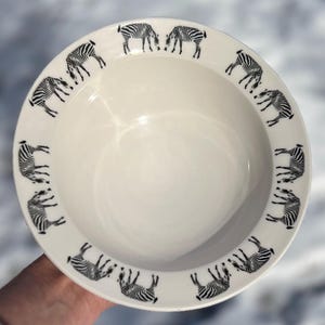 May include: White ceramic bowl with a black zebra pattern around the rim. The bowl has a wide, shallow design, and the zebra illustrations are in a repeating pattern. The bowl is held by a hand.