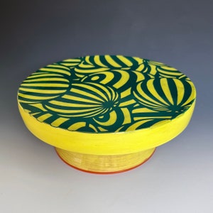 May include: A yellow ceramic cake stand with a green and yellow geometric pattern. The stand has a round top and a cylindrical base.