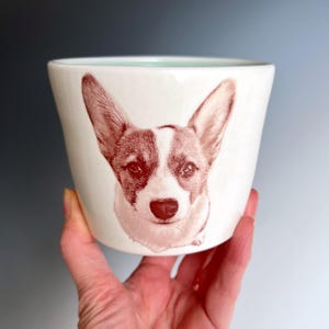 Puede incluir: Una taza de cerámica blanca con una imagen roja y blanca de un perro corgi impresa en la parte delantera.