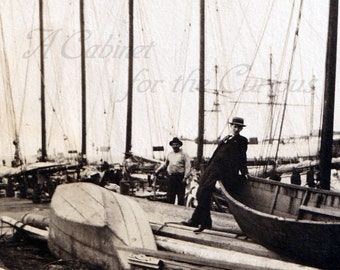 May include: A black and white photo of a group of boats docked at a pier. The boats are all wooden and have tall masts. There are two men standing on the pier, one of whom is wearing a hat. The boats are surrounded by wooden planks.