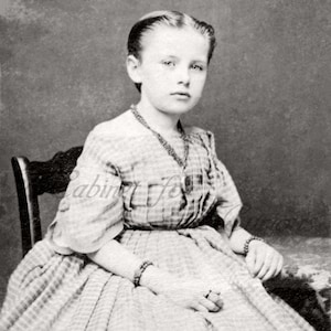 May include: A young girl wearing a gingham dress with a white collar and a necklace. She is sitting on a chair with her hands in her lap.