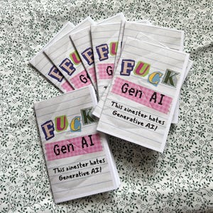 Puede incluir: Una pila de fanzines con el texto "FUCK Gen AI" en letras coloridas y cuadradas. La frase "This zinester hates Generative AI!" también está impresa en la portada. Los fanzines están sobre papel rayado con una cinta rosa.