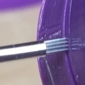 May include: Close-up of a silver metal needle with a purple plastic object in the background. The needle is inserted into the plastic object, and the tip of the needle is visible.