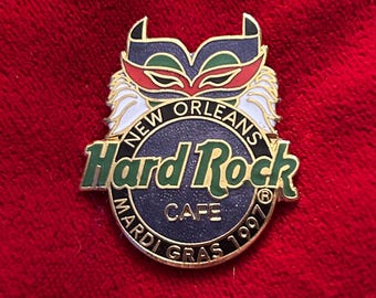 May include: A Hard Rock Cafe pin featuring a Mardi Gras mask design. The pin has a dark blue background with the words "New Orleans" and "Hard Rock Cafe" in green lettering. The pin also says "Mardi Gras 1997".