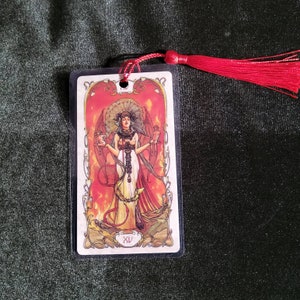 May include: A tarot card bookmark featuring a detailed illustration of a woman with wings, holding objects, set against a fiery red background. The bookmark has a red tassel attached. The card is encased in a clear plastic sleeve.