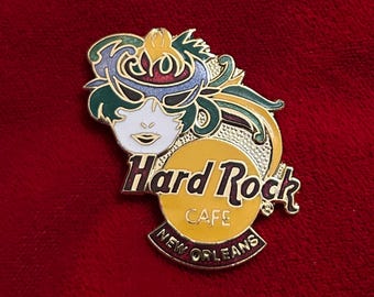 May include: A Hard Rock Cafe pin from New Orleans, featuring a Mardi Gras mask design. The pin has a yellow circle with the cafe's logo and the city's name. The mask is white, green, blue, and gold.