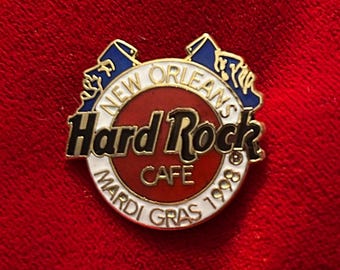 May include: A Hard Rock Cafe pin from New Orleans, featuring a circular design with the words "Hard Rock Cafe" in black and white, surrounded by a red and white border. The pin also includes "Mardi Gras 1998" and blue accents.