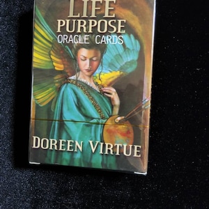 May include: A deck of oracle cards titled "Life Purpose Oracle Cards" by Doreen Virtue. The box features a painting of a woman with wings, holding a paintbrush and palette.