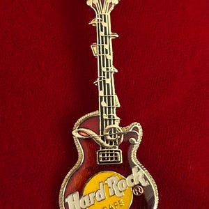 May include: A guitar-shaped pin with a gold border and a red and brown body. The pin features the Hard Rock Cafe logo and the city name "Nagoya" in white lettering on a yellow circle.