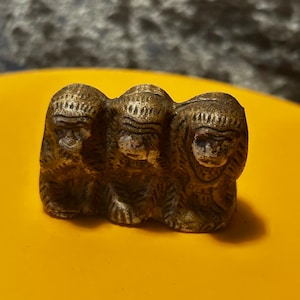 May include: Three small, brown, ceramic monkeys with closed eyes, sitting side-by-side. The monkeys are facing the same direction, with the monkey in the back looking over the shoulder of the monkey in front of it.