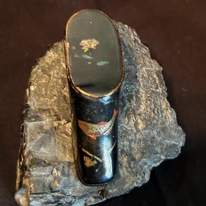 May include: A small, black lacquered box with gold and red painted details. The box is decorated with a floral design and a fish. The box is sitting on a piece of gray rock.