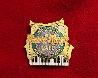 May include: A gold-colored Hard Rock Cafe pin from New Orleans, featuring a green and silver circular logo with the text "New Orleans Hard Rock Cafe" and "Mardi Gras 1992." The pin includes a piano keyboard and musical instrument accents.