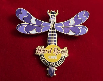 May include: A Hard Rock Cafe New Orleans dragonfly pin. The pin features a purple and white dragonfly design with gold accents. The Hard Rock Cafe logo is in the center on a yellow background.