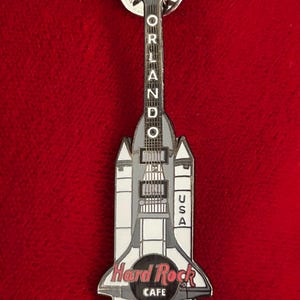 May include: A Hard Rock Cafe pin in the shape of a guitar and space shuttle. The pin is silver, white, and black, with the words "Orlando" and "USA" on it. The Hard Rock Cafe logo is also visible.