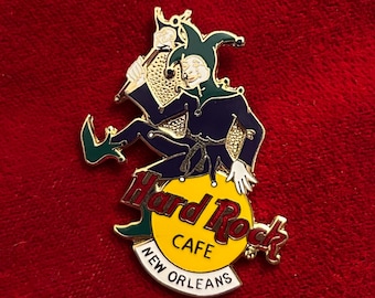 May include: Hard Rock Cafe New Orleans pin featuring a jester in a blue and green outfit, playing a horn. The pin has a yellow circle with the Hard Rock Cafe logo and the city name.