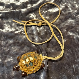 May include: A brown leather cord necklace with a ceramic turtle pendant. The turtle is decorated with white and blue glaze.