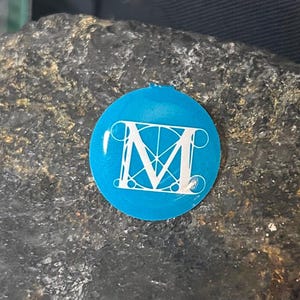 May include: A round, bright blue sticker with a white, stylized letter "M" in the center. The sticker is set against a dark, textured background, creating a contrast between the vibrant blue and the darker tones.