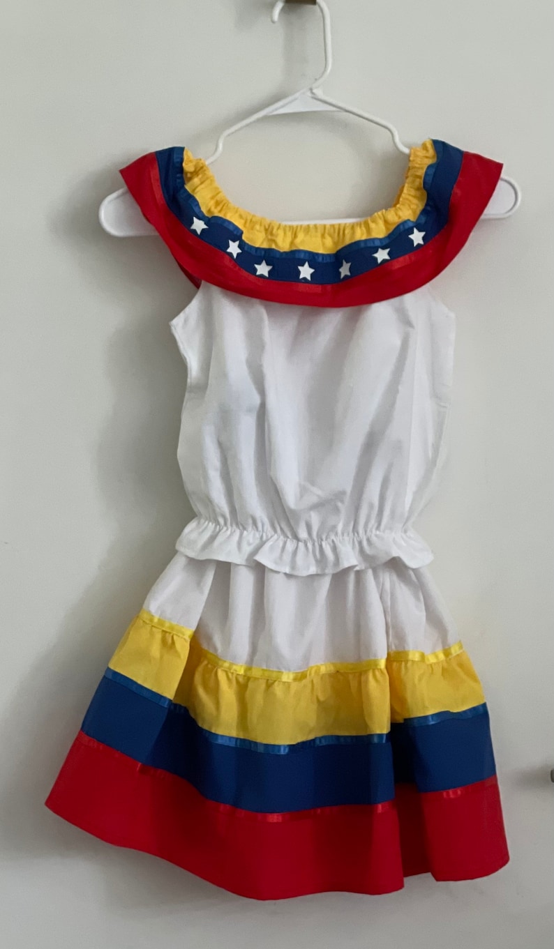 May include: A white sleeveless dress with a ruffled neckline featuring a blue and yellow ribbon with white stars. The skirt has yellow, blue, and red stripes.
