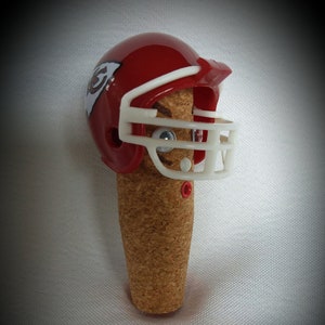 May include: A red and white football helmet with a cork body and the word "CHIEFS" on a black ribbon. The helmet has a white face mask and a red and white logo on the side.