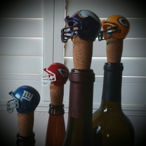 May include: Four wine bottle stoppers shaped like football helmets. The helmets are in the colors of the New York Giants, Kansas City Chiefs, Minnesota Vikings, and Philadelphia Eagles.