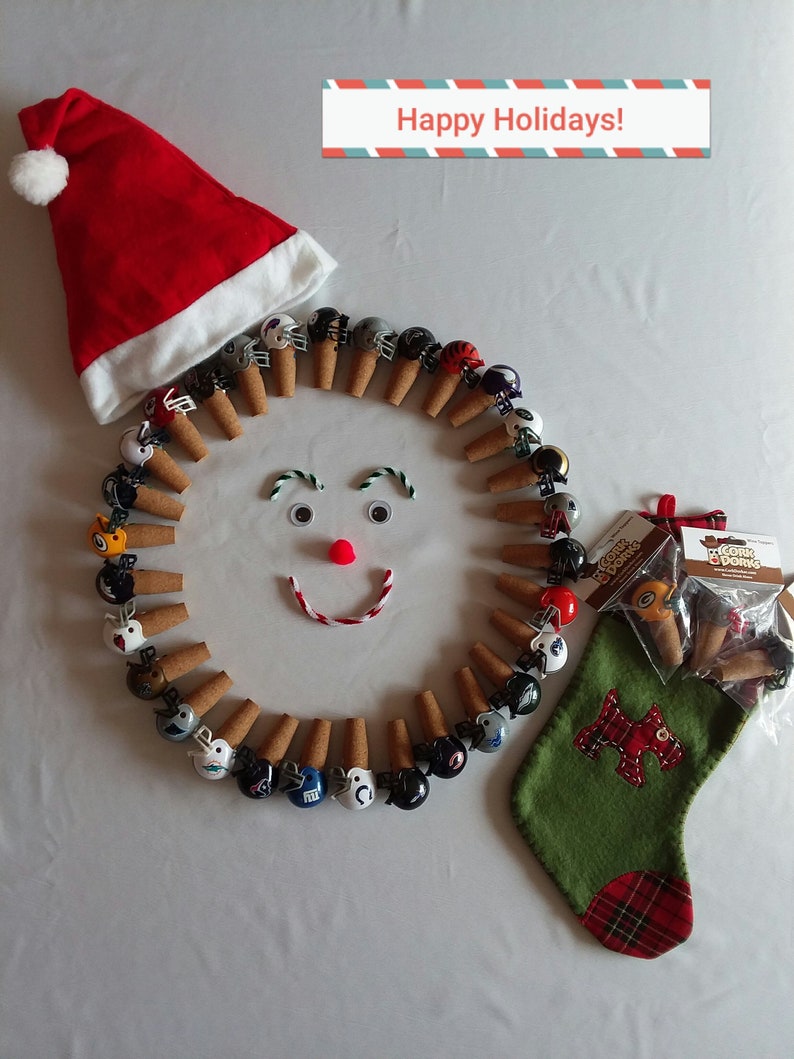 May include: A festive holiday scene with a red Santa hat, a green stocking with a red plaid cuff, and a circle of miniature football helmets on cork stoppers. The helmets are in various team colors and logos. The text "Happy Holidays!" is printed on a red and blue striped banner.