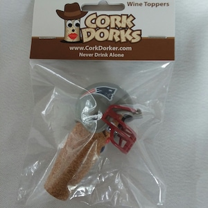 May include: A wine bottle stopper featuring a miniature football helmet in the colors of the New England Patriots. The helmet is gray with a red face mask and a blue and white logo. The stopper is made of cork and is packaged in a clear plastic bag with a white card that reads "Cork Dorks" and "Never Drink Alone".
