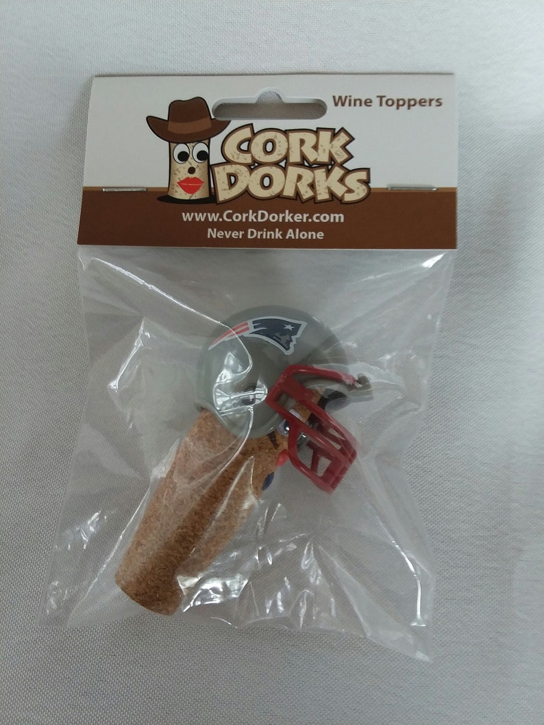 May include: A wine bottle stopper featuring a miniature football helmet in the colors of the New England Patriots. The helmet is gray with a red face mask and a blue and white logo. The stopper is made of cork and is packaged in a clear plastic bag with a white card that reads "Cork Dorks" and "Never Drink Alone".