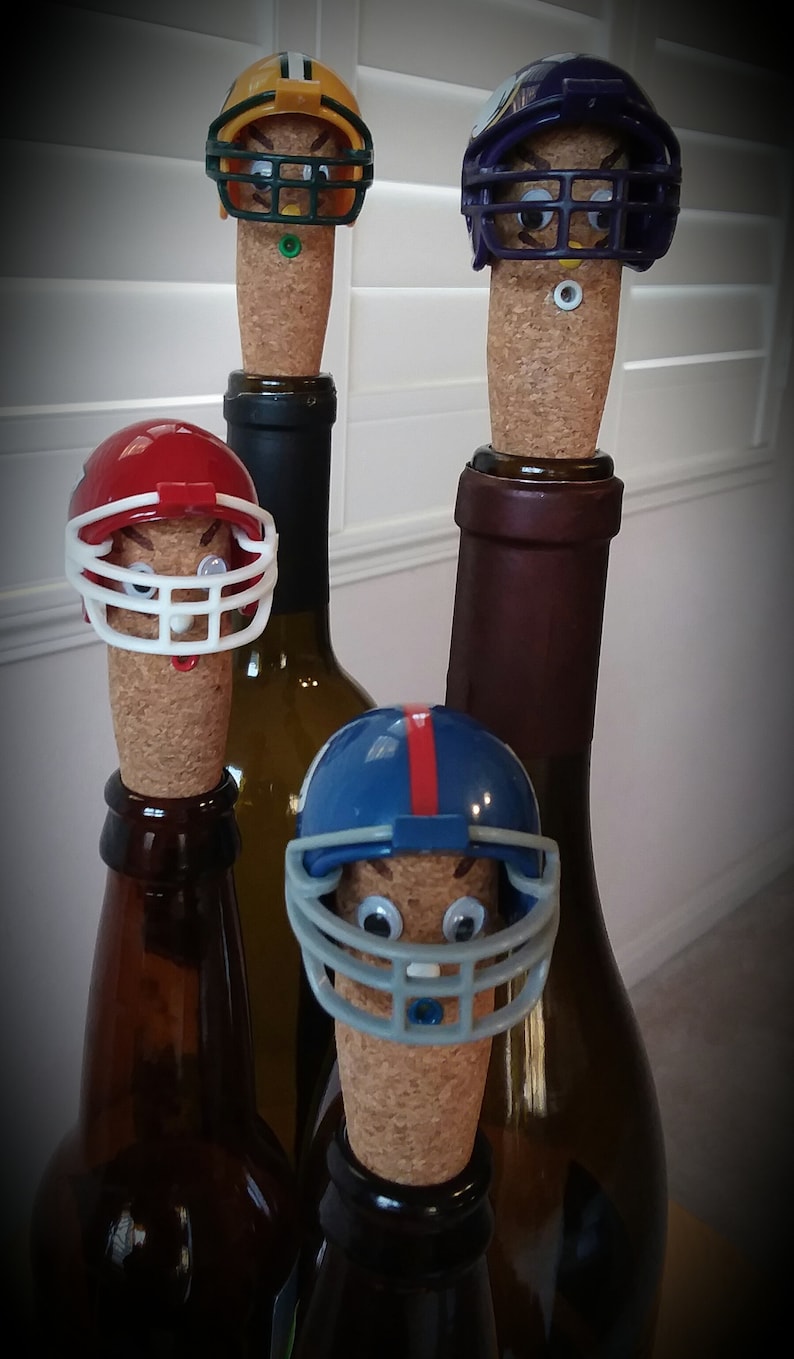 May include: Four wine bottle stoppers shaped like football helmets. The helmets are in the colors of different NFL teams: green and yellow, purple, red, and blue. The stoppers are on top of wine bottles.