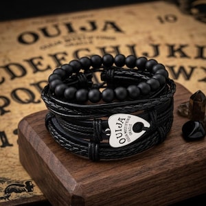 May include: A stack of black bracelets with a Ouija planchette charm. The bracelets include beaded and braided leather styles. The charm is silver with the word "Ouija" and "Mystifying Oracle" printed on it. The bracelets are displayed on a wooden surface.