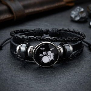 May include: Black leather bracelet with a braided band and a round, silver-toned centerpiece. The centerpiece features a white illustration of a drum set on a black background. The bracelet has adjustable cords.